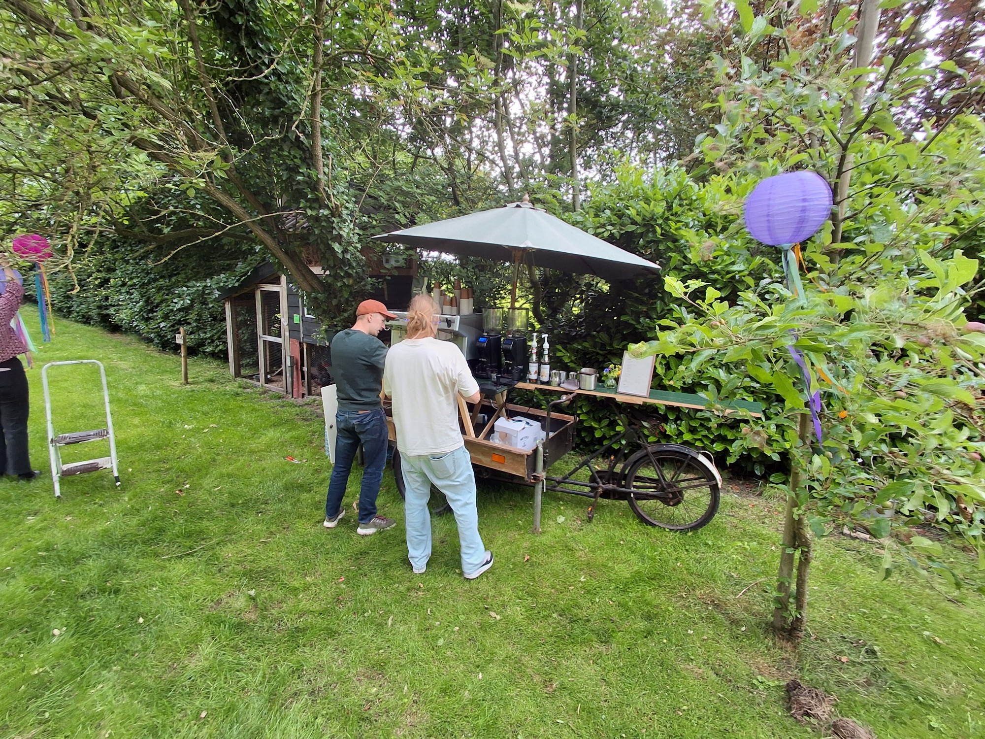 Twee mannen bij de mobiele koffiekar van Absoluukt onder een parasol in de tuin